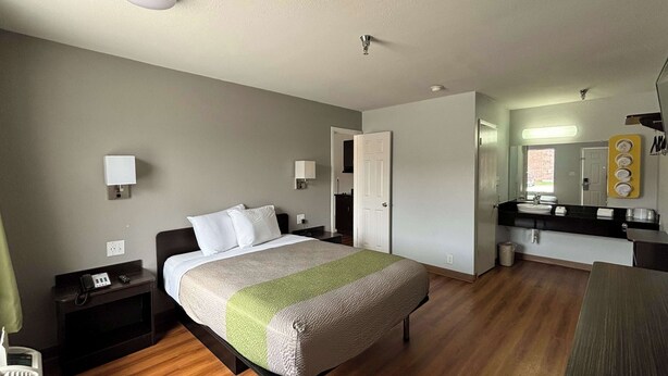 Una habitación de hotel con cama, mesita de noche, lámpara y baño con lavabo y espejo.