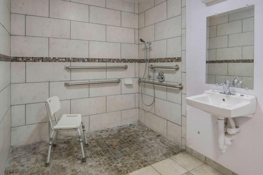 A bathroom with white tiles, a white sink, a mirror, and a wheelchair-accessible shower.