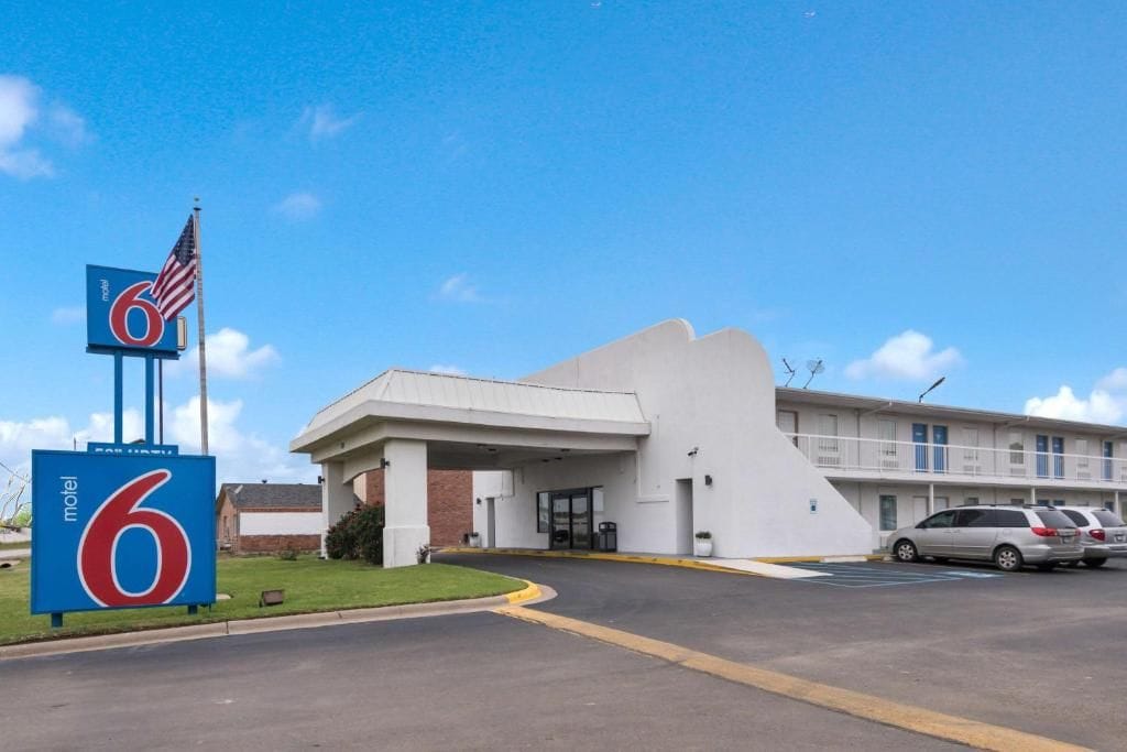 A motel with a large '6' sign, a parking lot, and a building with a sloped roof.