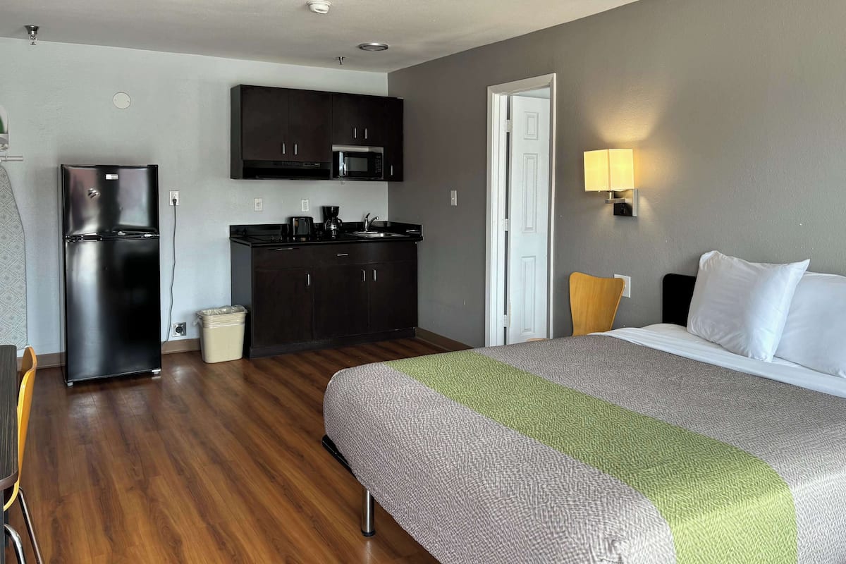 A hotel room with a bed, a small kitchenette with a refrigerator and microwave, a chair, and a bedside table with a lamp.