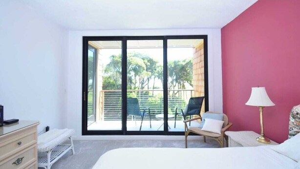 A bedroom with a large sliding glass door leading to a balcony, a bed, a dresser, a chair, and a lamp.