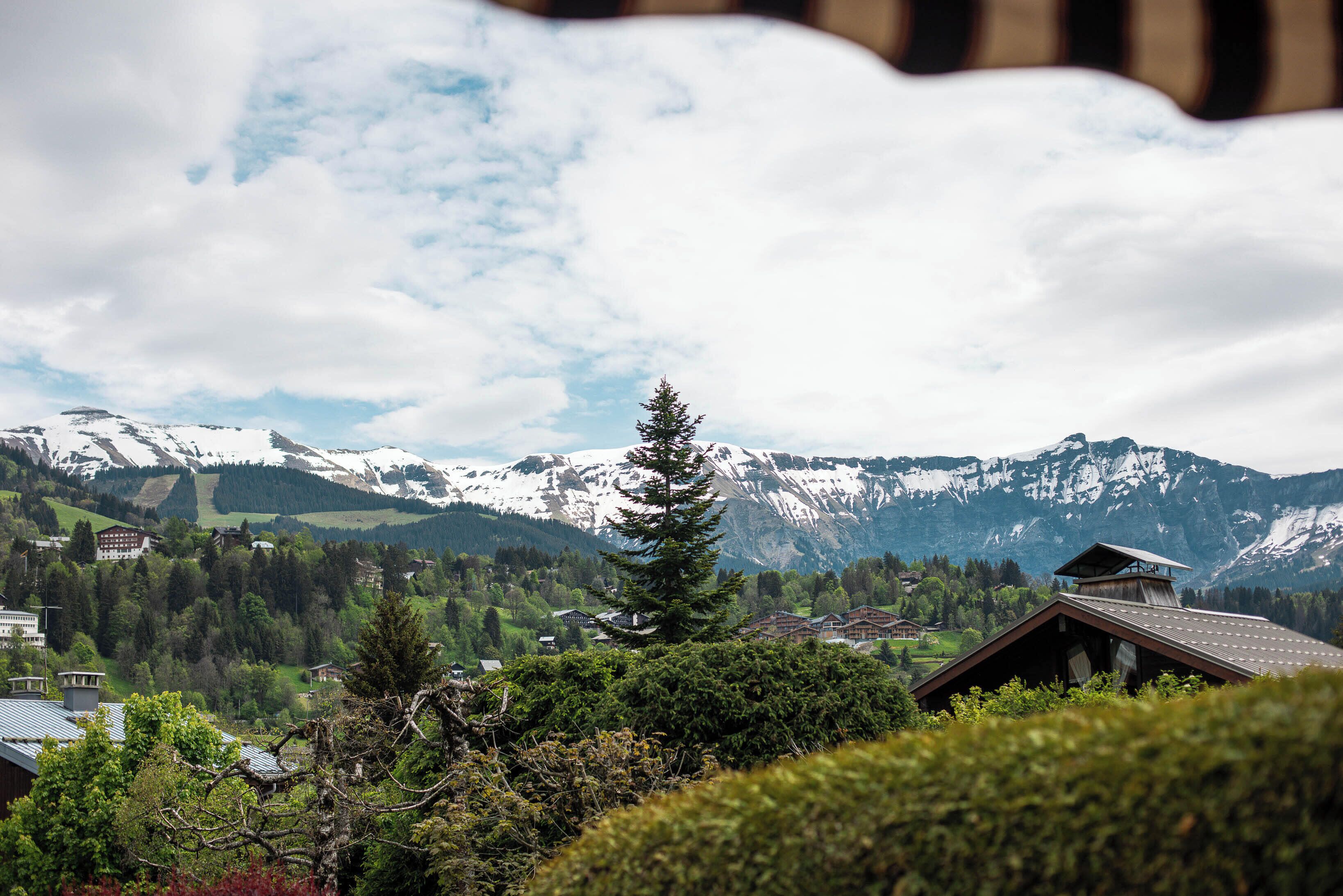 Un paysage montagneux avec des sommets enneigés, un grand arbre à feuilles persistantes qui se distingue, et des bâtiments traditionnels aux toits inclinés.