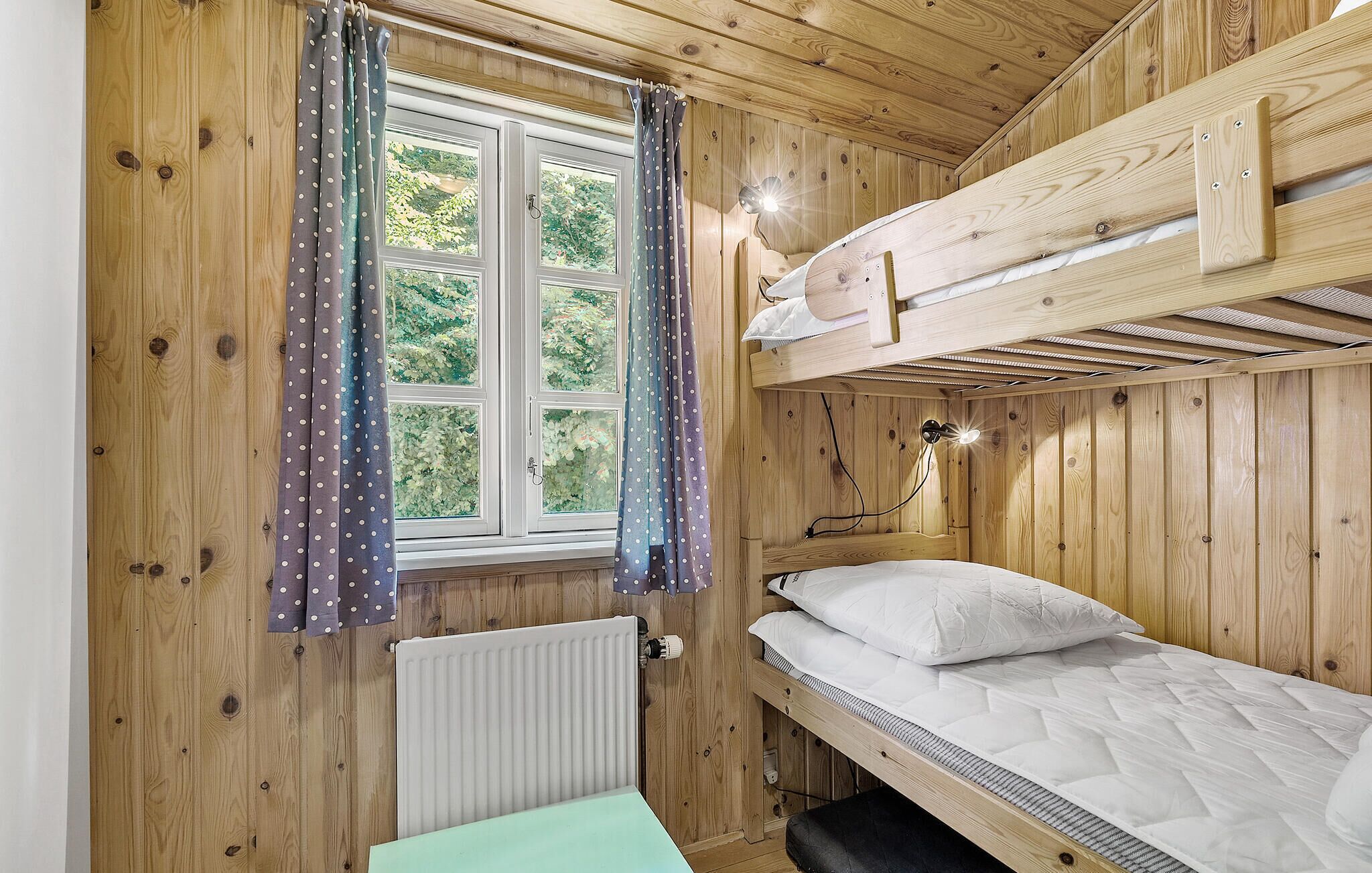 Een houten kamer met een stapelbed, een raam met gordijnen, een radiator en een bureau.