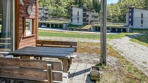 Una cabaña de madera con un área de patio que incluye mesas y bancos de madera, con vista a un complejo residencial de arquitectura moderna y un sendero de piedra.