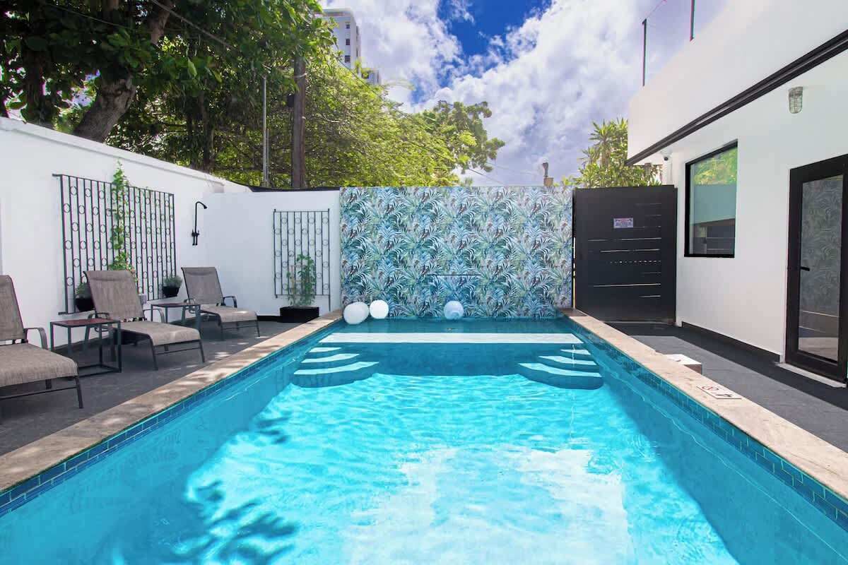Una piscina con sillas de descanso, un muro decorativo y un edificio con puerta y ventanas.