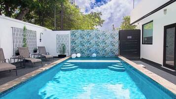 Una piscina con sillas de descanso, un muro decorativo y un edificio con puerta y ventanas.