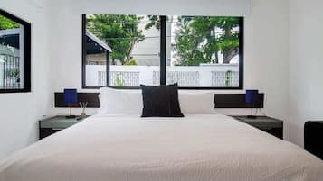 Una habitación de hotel con una cama grande, dos mesitas de noche con lámparas, un ventanal con vistas a árboles y un balcón, y paredes blancas.