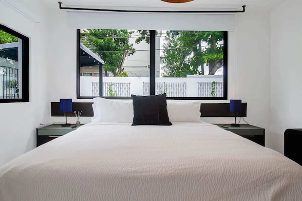 A hotel room with a large bed, two bedside tables with lamps, a window with a view of trees and a balcony, and white walls.