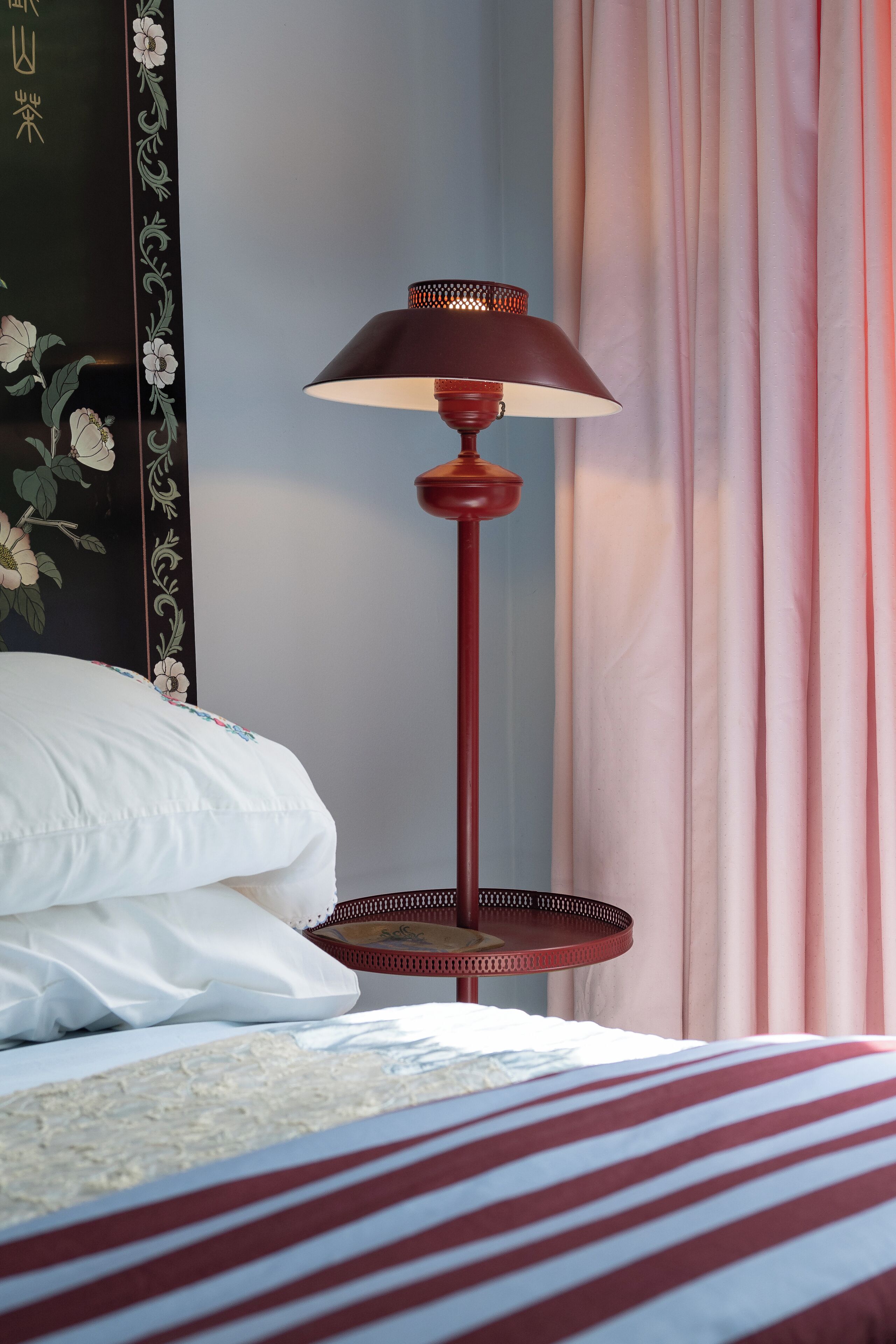 Un lit avec une literie blanche, une lampe de sol rouge et un mur orné d’œuvres d’art florales.