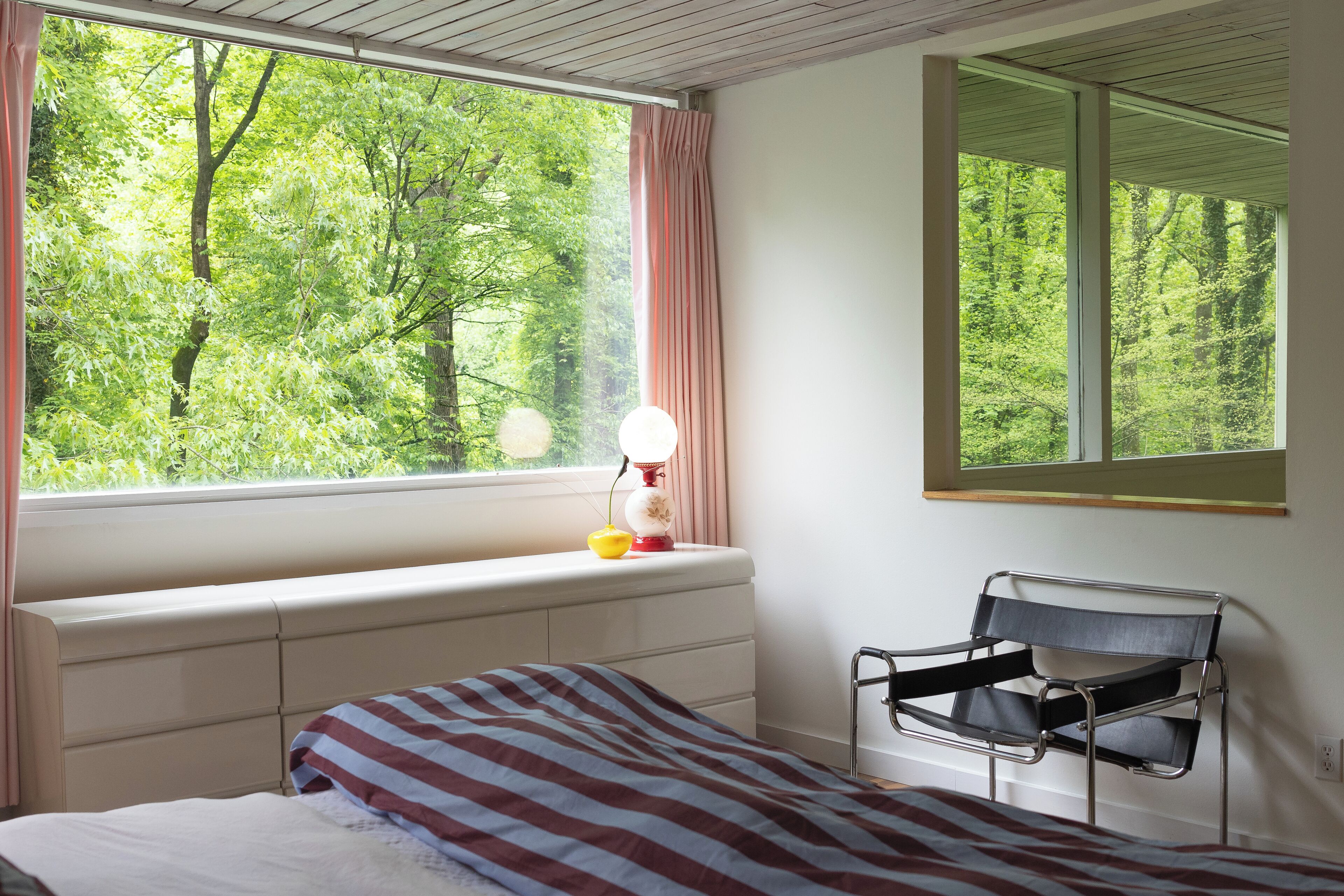Een slaapkamer met een raam, een bed, een stoel en een nachtkastje met een lamp.