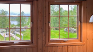 Zwei holzrahmenverziarte Fenster mit Metallgitter, mit Blick auf eine grüne Landschaft mit Felsen und einem Gewässer.