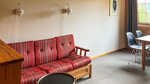 Ein Zimmer mit einer rot gemusterten Couch, einem kleinen runden Tisch, zwei Wandlampen, einem gerahmten Bild an der Wand und einem Essbereich mit Stühlen und Tisch.