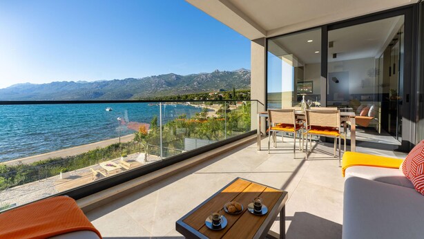 A balcony with a view of the sea and mountains, a dining area with chairs and a table, and a sofa with cushions.