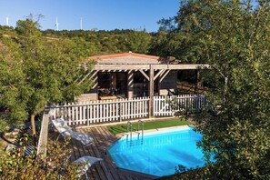 A swimming pool with a wooden deck and a covered seating area, surrounded by trees and a white picket fence.