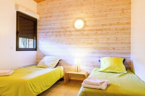 A room with two beds, each with a green bedspread and pillows, a small bedside table with a lamp, and a window with a closed blind.