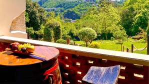 Un balcon avec une table où sont disposés des fruits, donnant sur un paysage verdoyant et luxuriant, avec des arbres et une vue lointaine sur une colline parsemée de bâtiments.