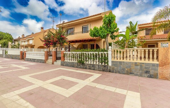 Zona residencial con un sendero pavimentado, entrada vigilada y un edificio con tejado de tejas y balcón.