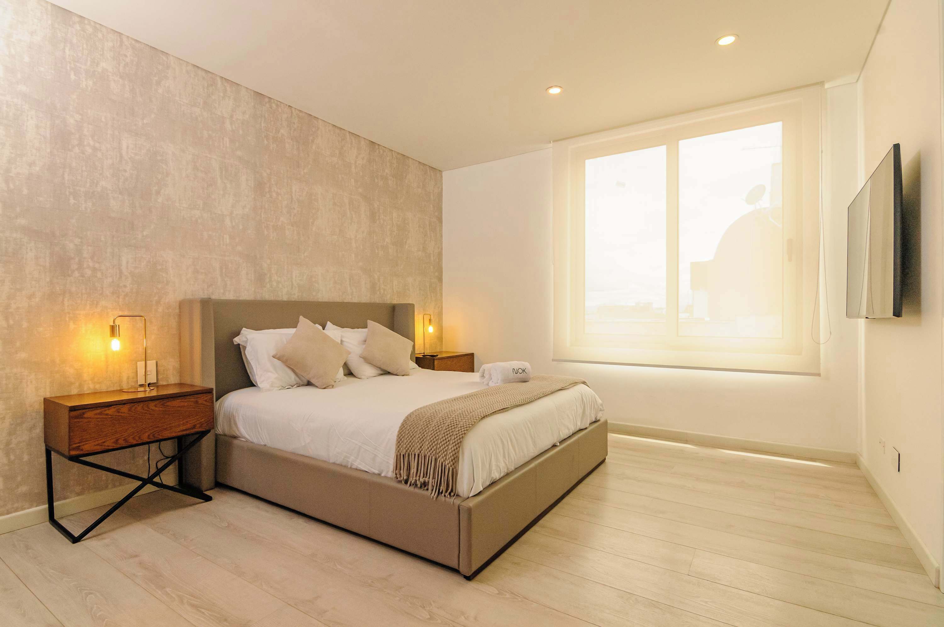 Une chambre moderne avec un grand lit, des tables de chevet avec des lampes, une fenêtre avec des stores et un téléviseur fixé au mur.