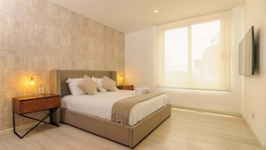 Une chambre moderne avec un grand lit, des tables de chevet avec des lampes, une fenêtre avec des stores et un téléviseur fixé au mur.