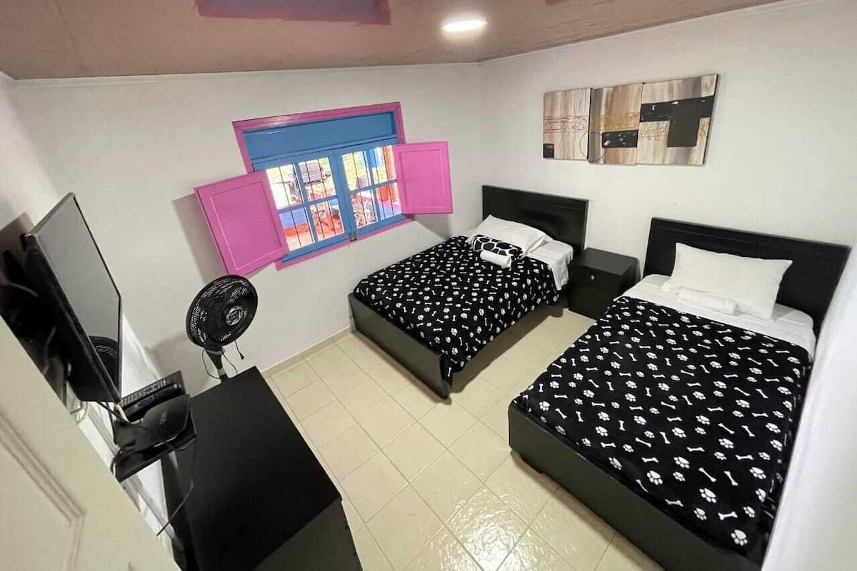 A bedroom with two beds, a desk with a computer, a fan, and a window with pink shutters.