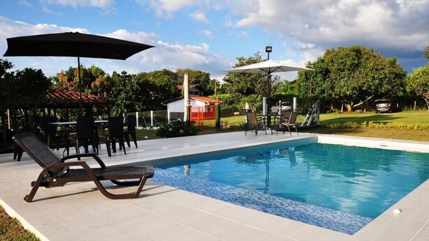 An outdoor swimming pool with lounge chairs and umbrellas, surrounded by a fenced garden and a small building. - finca el arrullo (Armenia)
