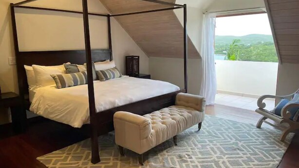 A four-poster bed with a bench, a patterned rug, and a view of the outdoors.