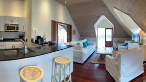 A modern kitchen with a central island, a dining area with a white table and chairs, and a living area with a sofa and a lamp.