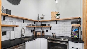 Una cocina moderna con armarios blancos, encimeras negras y electrodomésticos de acero inoxidable. Hay repisas de madera sobre las encimeras y un reloj en la pared.