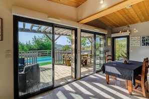 Een kamer met een schuifglazen deur die uitkomt op een terras met zwembad, houten meubels en een tafel met een fruitschaal.