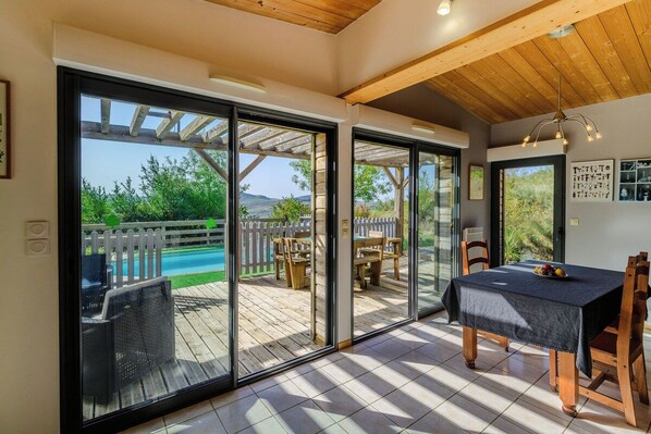 A room with a sliding glass door leading to an outdoor patio with a pool, wooden furniture, and a table with a fruit bowl.