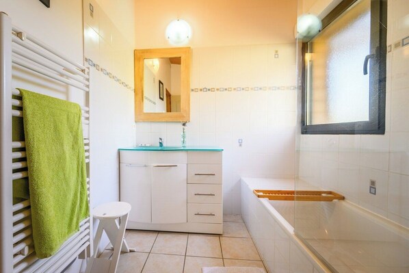 Un baño con pared de azulejos blancos, una toalla verde sobre un perchero blanco, un mueble blanco con un espejo y una bañera con borde de madera.