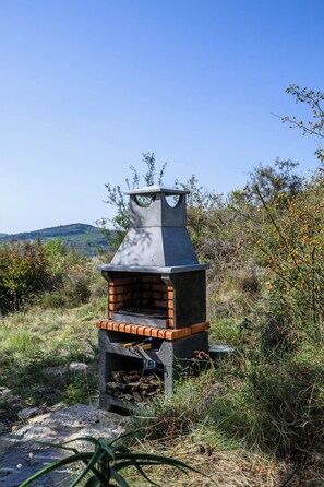 Una parrilla de ladrillo para barbacoa al aire libre con chimenea, rodeada de vegetación natural y colinas al fondo.