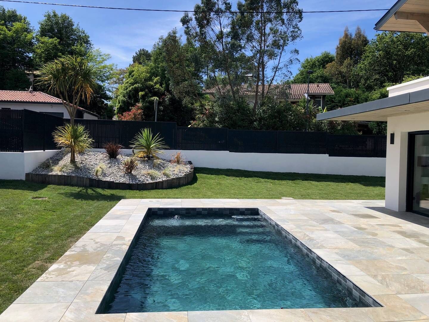 A rectangular swimming pool with a tiled border, surrounded by a well-maintained lawn and a garden with various plants and a palm tree.