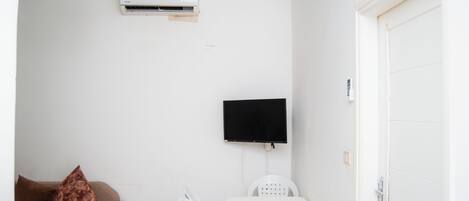 A room with a bed, a TV mounted on the wall, an air conditioner, a sofa with a cushion, and a door.
