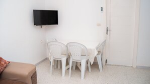 Una habitación con una mesa blanca, cuatro sillas blancas, un televisor negro montado en la pared, una puerta blanca y un sofá marrón.