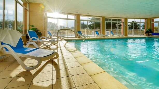 Une piscine intérieure à l’eau d’un bleu cristallin, entourée de grandes fenêtres baignées de lumière et de chaises longues.