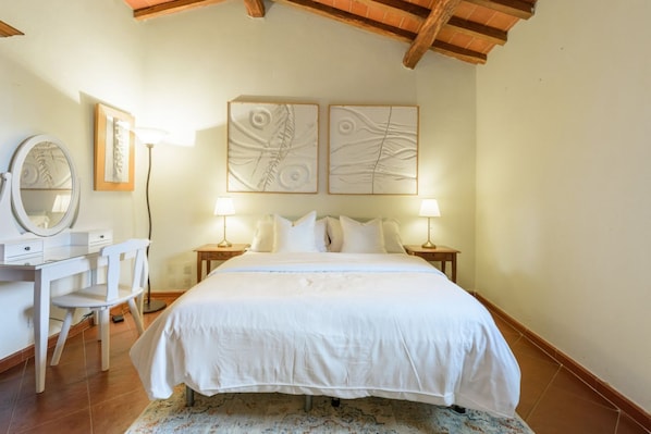 Une chambre à coucher comprenant un lit, des tables de chevet, un bureau avec une chaise, un miroir et des œuvres d’art accrochées au mur.
