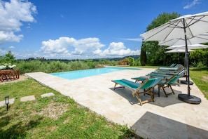Piscina com espreguiçadeiras e guarda-sóis, cercada por grama e árvores.