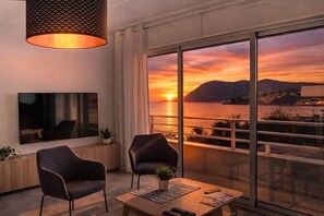 Una habitación moderna con televisión, dos sillones, una mesita de centro y un amplio ventanal con vistas al atardecer.