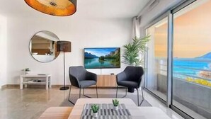 Een moderne woonkamer met een groot raam, een flatscreen televisie, twee stoelen, een ronde spiegel en een kleine tafel met een plant.
