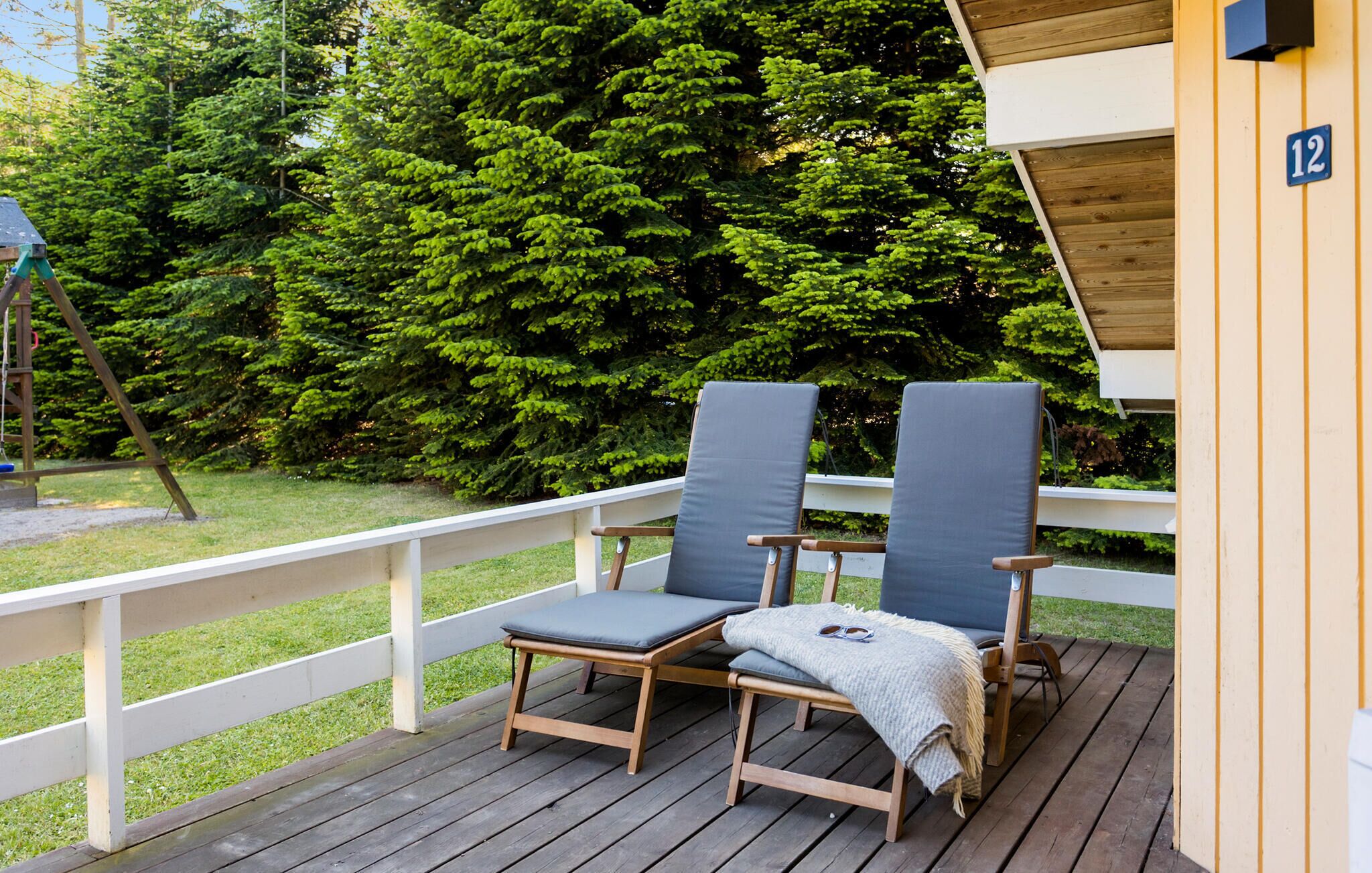 Eine Holzterrasse mit zwei Liegestühlen und einem kleinen Tisch, umgeben von einem weißen Geländer und dichtem Grün der Bäume.