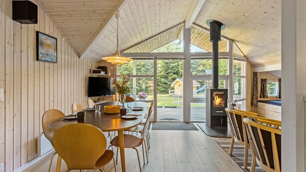 A cozy cabin interior with a dining area, a fireplace, and large windows offering a view of the outdoors. - Stunning home in Tranekær with sauna (Tranekær)