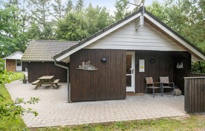 Une cabane en bois avec un toit en tuiles, une table de pique-nique et deux chaises à l’extérieur.