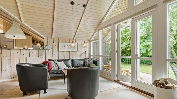 Una sala de estar acogedora con un sofá de cuero negro, dos sillones, techo de madera y ventanas amplias que ofrecen vistas al exterior.
