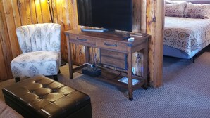 Ein gemütliches Zimmer mit einer Holzwand, einem Flachbildfernseher auf einem Holzbodenständer, einem gemusterten Sessel, einem Sofa mit floralen Kissen und einem Lederetageere.
