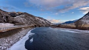 Ein Fluss fließt durch eine gebirgige Landschaft, mit schneebedeckten Hügeln und einer Brücke auf der linken Seite.