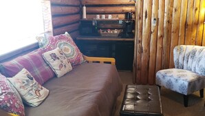 Ein gemütliches Blockhauszimmer mit einem Bett, einem Sessel, einem kleinen Tisch und einer Küchenzeile.