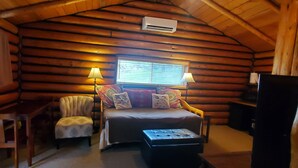 A log cabin room with a bed, two chairs, a table, and a window with blinds.
