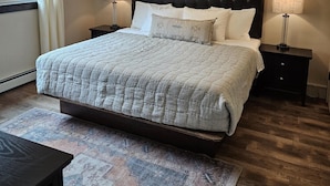 Un dormitorio con una cama grande, dos mesitas de noche, una lámpara colgante, un cuadro en la pared y una ventana con cortinas.