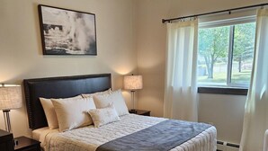 Une chambre à coucher avec un grand lit, deux tables de chevet, une commode, une fenêtre avec des rideaux et un tableau encadré au mur.